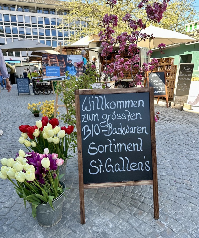 Kreidetafel am Marktplatz: Willkommen zum grössten Bio-Backwaren-Sortiment St. Gallens, daneben Tulpen in Eimern